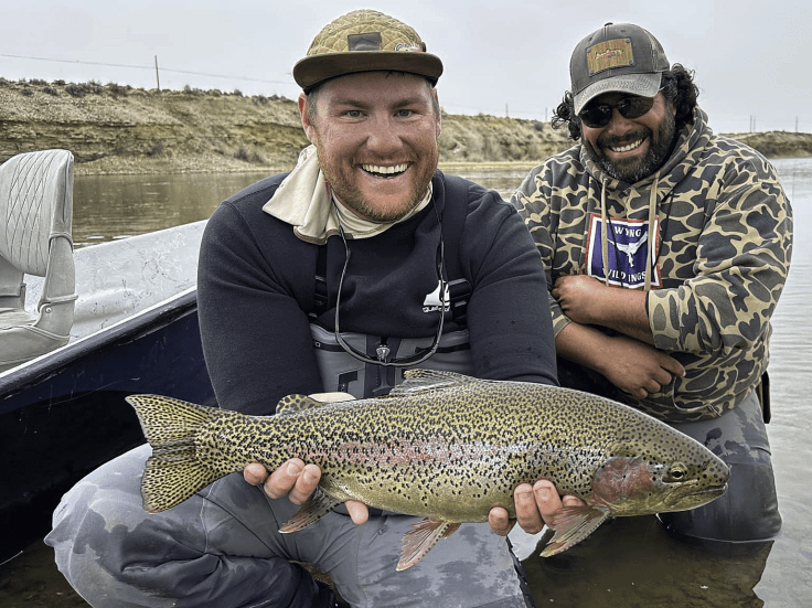 A smiling angler holding a large rainbow trout while seated in a boat, with a fellow angler in the background sharing a joyous moment.