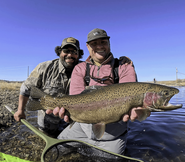 Two anglers stand in shallow water, smiling as they hold a large rainbow trout. One angler has a mustache and wears sunglasses and a camouflage shirt, while the other wears a cap and a pink long-sleeve shirt. A fishing net is visible in the foreground.